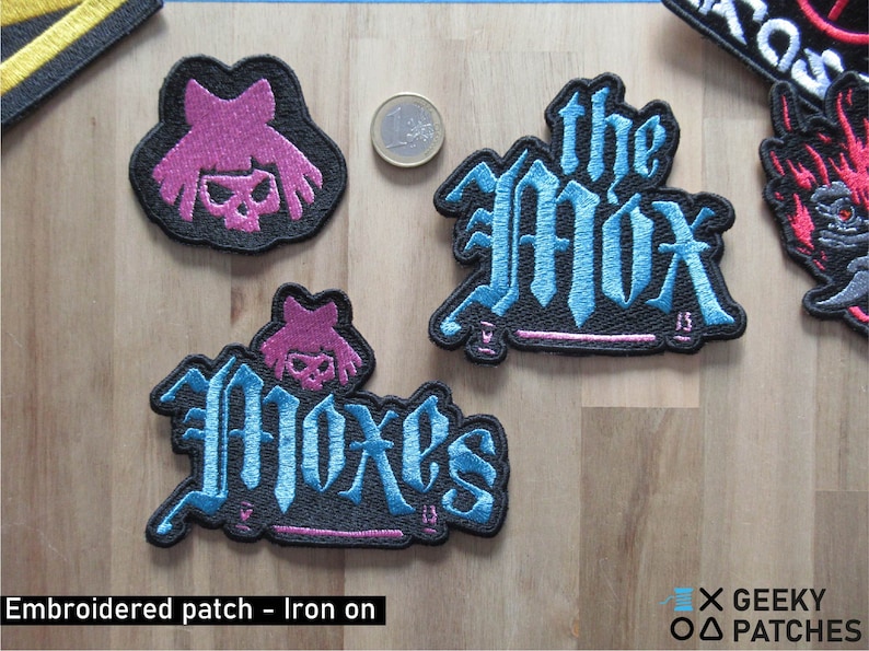 May include: Three embroidered iron-on patches featuring the band "The Mox" in blue and pink lettering on a black background. The patches are on a wooden surface.