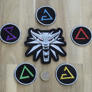 Puede incluir: Seis parches bordados con la cabeza de un lobo, un triángulo morado, un triángulo azul, un triángulo rojo, un triángulo verde y un triángulo amarillo. Los parches están dispuestos en círculo alrededor del parche de la cabeza de lobo.