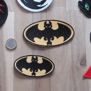 Könnte beinhalten: Zwei gestickte Aufnäher mit einer schwarzen Silhouette einer Fledermaus mit gelben Akzenten. Die Aufnäher sind oval geformt und haben einen gelben Umriss.
