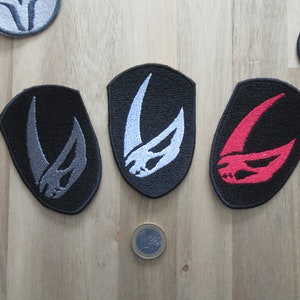 May include: Three embroidered patches with a black background and a white, grey, and red Mandalorian symbol. The patches are on a wooden surface.