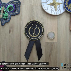 May include: A black and gold embroidered patch with a geometric design. The patch is attached to a black ribbon and has the text "Iron On OR Sew On" and "6.5 x 6.5 cm (12 cm with te ribbon) / 2.56 x 2.56 inch (5 inch)" printed below it.