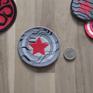 Könnte beinhalten: Ein grauer und schwarzer gestickter Aufnäher mit einem roten Stern in der Mitte. Der Aufnäher ist rund und hat ein Design, das einem Schild ähnelt.
