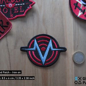Puede incluir: Parche bordado termoadhesivo con una V estilizada en negro y azul dentro de un círculo rojo con anillos concéntricos. El parche mide 8,5 cm por 6 cm.