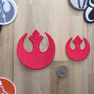 Peut inclure: Deux écussons rouges brodés avec le symbole de l'Alliance Rebelle, un grand et un petit. Les écussons sont circulaires avec un emblème stylisé en forme d'oiseau. D'autres écussons sont visibles en arrière-plan.