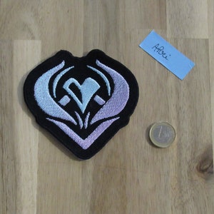 Puede incluir: Un parche bordado negro con un diseño en forma de corazón. El corazón está bordeado de blanco y relleno con un degradado de azul y morado. El parche es aproximadamente del tamaño de una moneda de 1 euro.