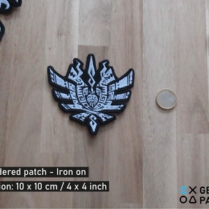 May include: Embroidered patch with a black border and white design, featuring a detailed emblem. The patch is 4 x 4 inches. Additional patches and a coin are visible on a wooden surface.