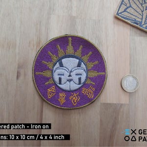 May include: Round, embroidered iron-on patch with a purple background and a sun-like design in gold and white. The patch measures 10 x 10 cm. The design includes a face and symbols. A coin is next to the patch.