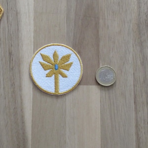 May include: A white circular patch with a yellow embroidered tree design. The tree has a blue gem in the center.