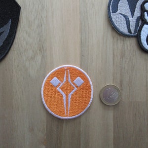 Peut inclure: Patch brodé orange et blanc avec un motif stylisé en forme de diamant avec un sommet pointu et deux petits diamants de chaque côté. Le patch est circulaire et a une bordure blanche.