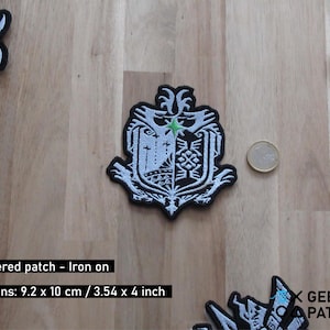 Könnte beinhalten: Weißer gestickter Bügel-Patch mit einem schwarzen und weißen Wappen-Design. Das Wappen zeigt einen Stern, ein Kreuz und ein stilisiertes Tier. Der Patch ist 9,2 cm x 10 cm groß.