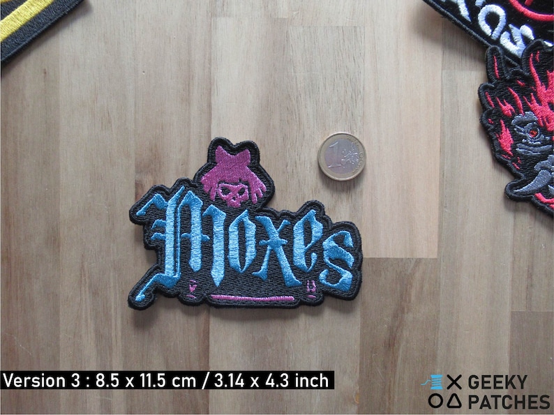 May include: Blue and pink embroidered patch with the word "Moxes" in a gothic font. The patch is 3.14 inches by 4.3 inches.