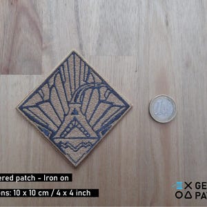 Puede incluir: Un parche de hierro bordado de color beige con un diseño geométrico azul. El parche mide 10 cm por 10 cm y presenta un diseño estilizado de rayos de sol.