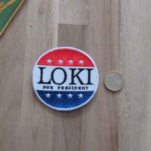 Może przedstawiać: Okrągła, haftowana naszywka z czerwono-niebieskim wzorem, z czarnym napisem "LOKI FOR PRESIDENT". Naszywka jest biała z pięcioma białymi gwiazdami u góry i na dole. Obok naszywki znajduje się moneta.