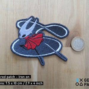 May include: Embroidered patch depicting a character with a white face, grey horns, and a red cape. The patch is dark blue with white trim and measures 7.5 x 10 cm. The text "Embroidered patch - Iron on" is visible.