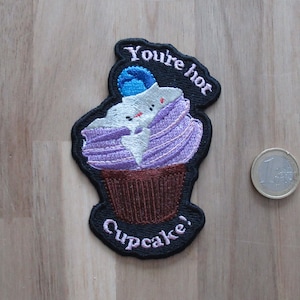 Puede incluir: Parche bordado con un diseño de cupcake y el texto "You're hot Cupcake!". La magdalena tiene glaseado morado, una cobertura azul y una base marrón, todo delineado en negro. Una moneda está al lado del parche.