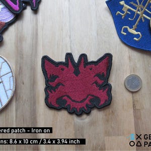 May include: Red embroidered iron-on patch featuring a two-headed creature with sharp teeth and claws. The patch is 3.4 inches by 3.94 inches.