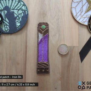 May include: Embroidered iron-on patch featuring a purple and brown geometric design with a green accent. The patch is rectangular, measuring 4.33 inches in length and 0.8 inches in width. Other patches and a coin are also visible.