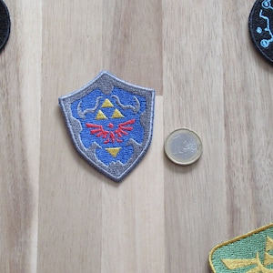 以下が含まれることがあります： ゼルダの伝説ビデオゲームシリーズのトライフォースシンボルを特徴とする青と灰色の刺繍パッチ。パッチは盾の形をしており、中央に赤い鳥のデザインがあります。