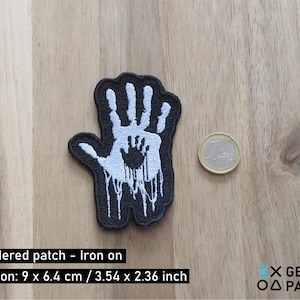 Könnte beinhalten: Weißer gestickter Bügel-Patch mit einer Hand, in der sich eine kleinere Hand befindet, die mit schwarzer Flüssigkeit tropft. Der Patch ist 9 cm x 6,4 cm groß.