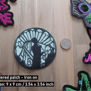 May include: An embroidered iron-on patch featuring a silhouette of a person standing in front of a tree with a white background. The patch is 9 inches by 9 inches and is surrounded by other patches on a wooden surface.