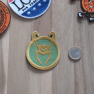 May include: A green and gold embroidered patch with a stylized image of a horned helmet and a single eye. The patch is circular and has a gold border.