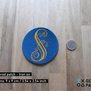 May include: A blue embroidered iron-on patch with a gold letter 'S' in a decorative font. The patch is 9 cm by 9 cm.