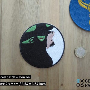 May include: Embroidered patch featuring a split design of a green-skinned witch and a white-haired woman. The patch is round, with dimensions of 3.54 x 3.54 inches. A blue patch with gold swirls and a coin are also visible.