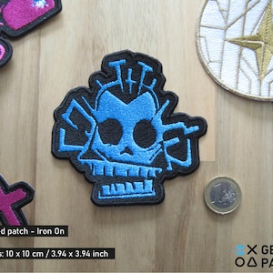 May include: Blue embroidered iron-on patch featuring a skull with the word "The" above it. The patch is 10 cm by 10 cm.