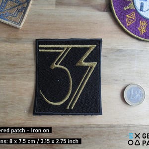 May include: Black embroidered patch with the number 37 in gold. The patch is square, measuring 3.15 x 2.75 inches. The text "Embroidered patch - Iron on" is visible. A coin is next to the patch.