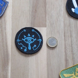 Könnte beinhalten: Ein schwarzer, kreisförmiger, gestickter Aufnäher mit einem stilisierten blauen Augendesign. Der Aufnäher hat einen blauen Rand mit kleinen Kreisen. Weitere Aufnäher mit ähnlichen Designs sind im Hintergrund sichtbar. Eine Münze ist ebenfalls vorhanden.