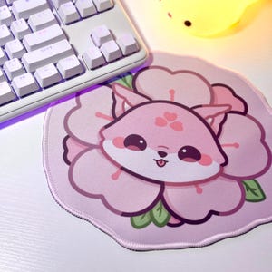 May include: A pink and white mousepad with a cartoon fox wearing a flower on its head. The fox has big eyes and a pink nose. The mousepad is round and has a stitched edge.