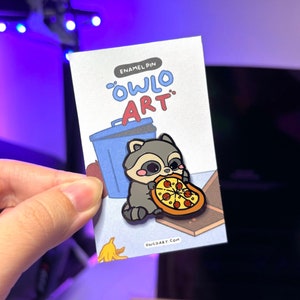 May include: Enamel pin featuring a cartoon raccoon holding a slice of pizza. The pin is on a card with the text "Enamel Pin "Owlo Art" owloart.com".