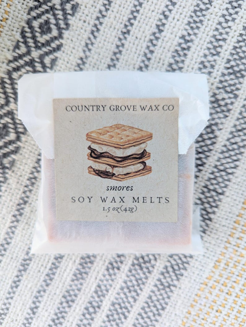 May include: A package of s'mores soy wax melts from Country Grove Wax Co. The label features a drawing of a s'more. The package is wrapped in white paper and rests on a patterned surface. The package weighs 1.5 oz (42g).