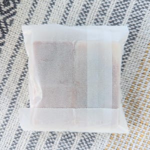May include: A square package wrapped in white paper. The contents are visible through the translucent paper, revealing a rectangular shape with a reddish-brown hue. The package is sealed with a strip of white tape.