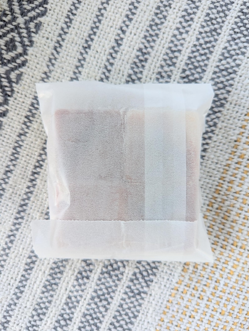 May include: A square package wrapped in white paper. The contents are visible through the translucent paper, revealing a rectangular shape with a reddish-brown hue. The package is sealed with a strip of white tape.
