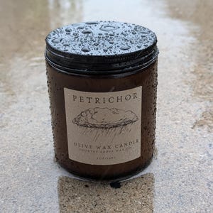 May include: A brown glass jar candle with a black lid, covered in water droplets. The label features the text PETRICHOR, an illustration of a rain cloud, and the words OLIVE WAX CANDLE, COUNTRY GROVE WAX CO, and 7OZ/198G in black ink.