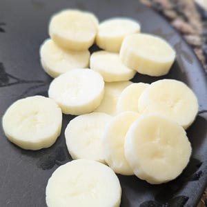 Op de afbeelding: Een zwart bord met een wit bloemenpatroon bevat een stapel gesneden banaan. De bananenplakjes zijn in een cirkelvormige vorm gerangschikt, waarbij de randen van de plakjes elkaar overlappen.