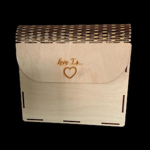 Könnte beinhalten: Eine hellfarbene Holzkiste mit gewölbtem Deckel und rechteckiger Basis. Der Deckel ist mit einem sich wiederholenden Muster und den Worten "Love Is..." mit einem Herzsymbol graviert. Die Kiste steht vor einem schwarzen Hintergrund.