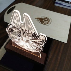 Könnte beinhalten: Ein klares Acryl-3D-Modell des Raumschiffs Millennium Falcon aus Star Wars, von innen beleuchtet. Das Modell wird auf einem Holzsockel mit einer kleinen Plakette präsentiert, auf der "Möge die Macht mit dir sein, immer" steht. Der Sockel befindet sich in einer schwarzen, mit Samt ausgekleideten Box.