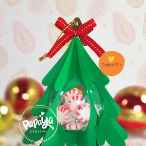 Puede incluir: Un adorno de árbol de Navidad de papel verde con una cúpula de plástico transparente que contiene bastones de caramelo. El adorno está decorado con una cinta roja y una cuerda dorada para colgar. El fondo es un patrón rosa y blanco de fruta de papaya.