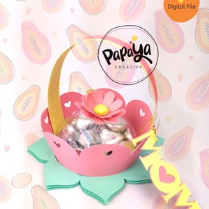 May include: A pink and green paper craft basket with a gold handle, filled with chocolate candies. The basket has a flower on top and a yellow tag that says "MOM" hanging from it. The background is a pink and white pattern of papaya fruit.