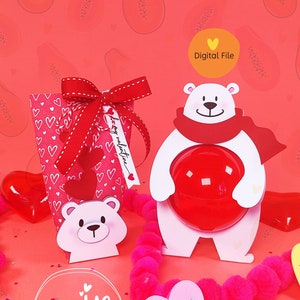 Puede incluir: Un archivo digital para un proyecto de manualidades de San Valentín con un oso polar blanco sosteniendo un corazón rojo. El oso lleva una bufanda roja. El proyecto también incluye una pequeña caja con un patrón de corazón y un corazón rosa con el texto "Be Mine".
