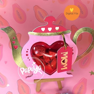 Puede incluir: Una caja en forma de tetera rosa y dorada con una ventana en forma de corazón llena de caramelos rojos. La caja está sobre un soporte de madera y tiene una etiqueta que dice "MAMÁ". El fondo es un patrón rosa y naranja de fruta de papaya.