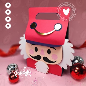 May include: A red and white paper nutcracker box with a gold handle, black mustache, and white beard. The box is decorated with a heart and the words "Digital File". Decorative Christmas ornaments are scattered around the box.