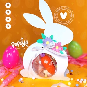 Puede incluir: Una caja de regalo blanca con forma de conejito de Pascua con una ventana redonda y transparente, llena de golosinas. La caja está decorada con flores de papel y una cinta. Un huevo rosa con lunares y un huevo verde están en el fondo.