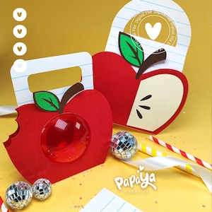 Puede incluir: Dos bolsas de regalo de papel con forma de manzana y asas con patrones de papel de cuaderno rayado. Una bolsa tiene una ventana circular transparente y un corte de mordisco, mientras que la otra muestra un diseño de manzana con hoja verde.
