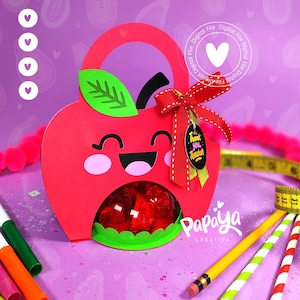 以下が含まれることがあります： 笑顔の顔、緑の葉、そして「先生ありがとう」と書かれた黄色のタグが付いた赤いリンゴの形のギフトバッグ。バッグは赤いリボンで飾られており、中のおやつが見える透明な窓があります。バッグはピンクのポンポンと鉛筆のある紫色の表面の上にあります。