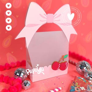 Könnte beinhalten: Eine rosa Geschenkbox mit einer großen Schleife oben und einem Kirschdesign. Die Box hat einen gewellten Rand und den Text "Papaya Creativa". Dekorative Discokugeln und ein Notizbuch sind ebenfalls im Bild.