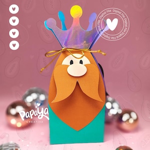 Puede incluir: Una caja de regalo de papel caprichosa con forma de rey, con una corona holográfica y barba naranja. La caja tiene una base turquesa y está atada con hilo dorado. El logotipo de Papaya Creativa es visible.