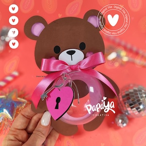 Puede incluir: Una etiqueta de regalo con forma de oso pardo con una cinta rosa y un adorno transparente. El adorno tiene una etiqueta rosa en forma de corazón con un diseño de llave y cerradura. El texto "Papaya Creativa" es visible.